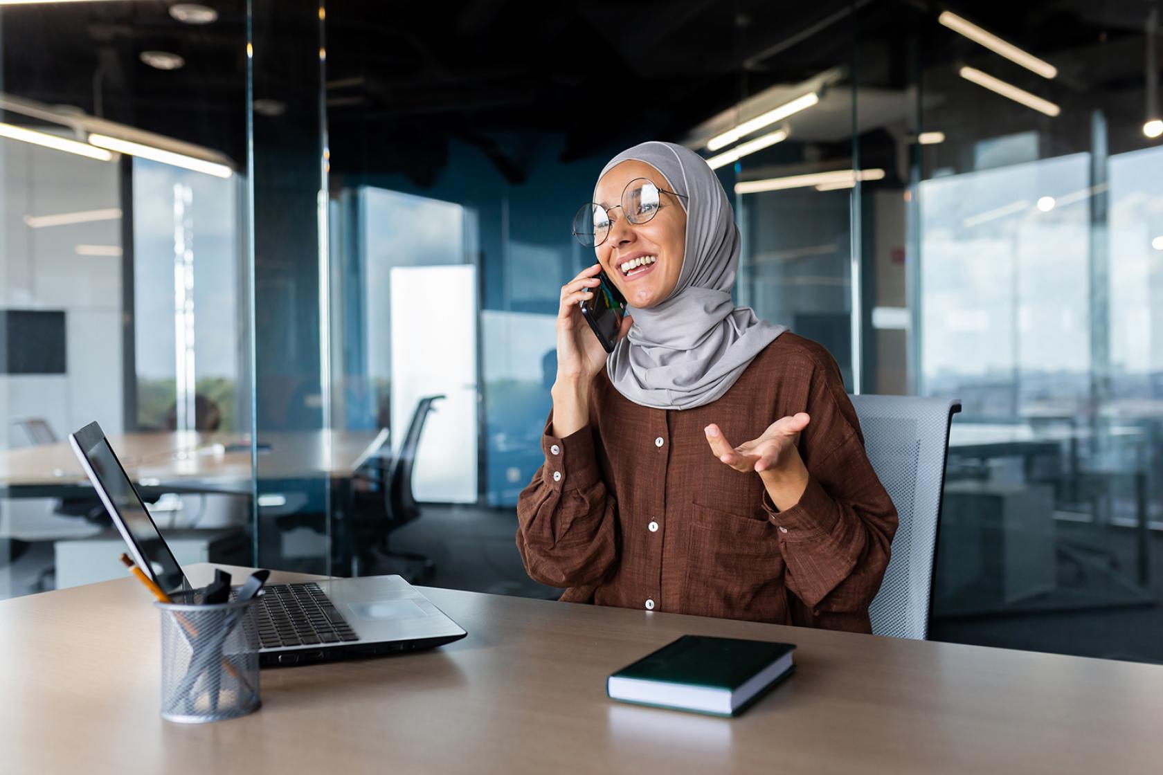 Vrouw met bril en hoofddoek is aan het bellen in een kantooromgeving, terwijl ze vrolijk kijkt en met haar hand gebaart.
